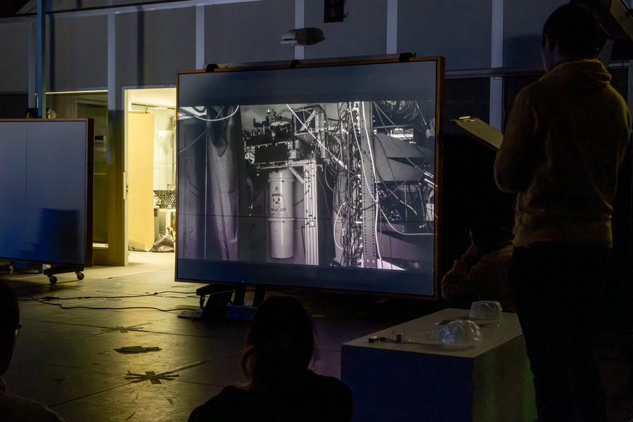In a darkened room, a person looking away from the camera views a grayscale photo of scientific equipment projected on a screen